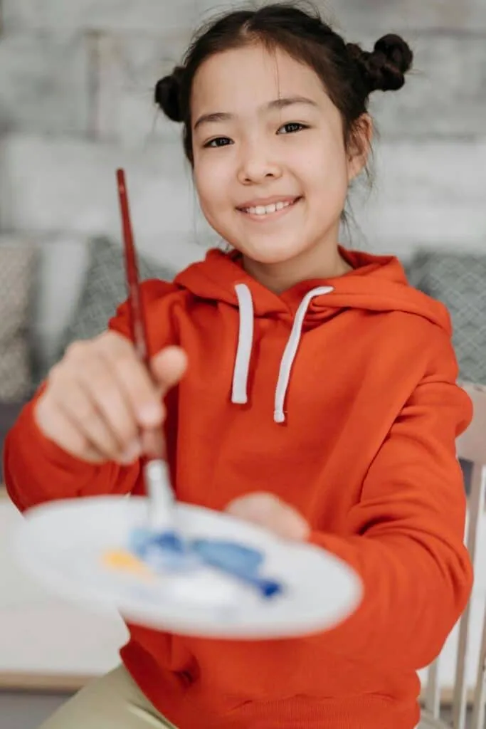 Girl in an orange hoodie holds a paintbrush and paint palette