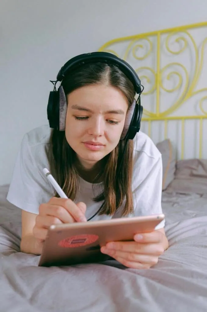Teen wearing headphones sketches a design on a tablet in bed