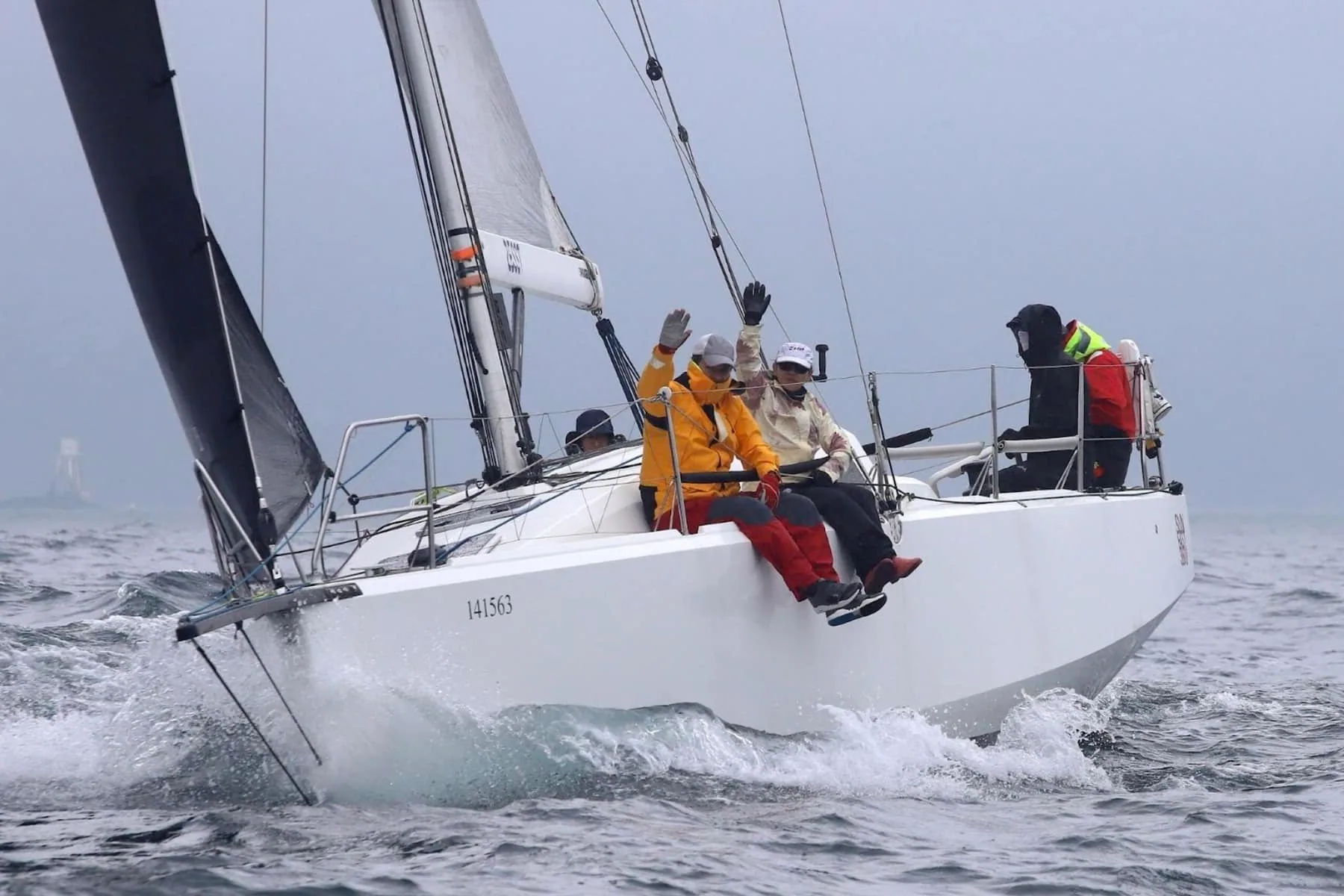 Offshore racing crew waves from the rail under a black mainsail