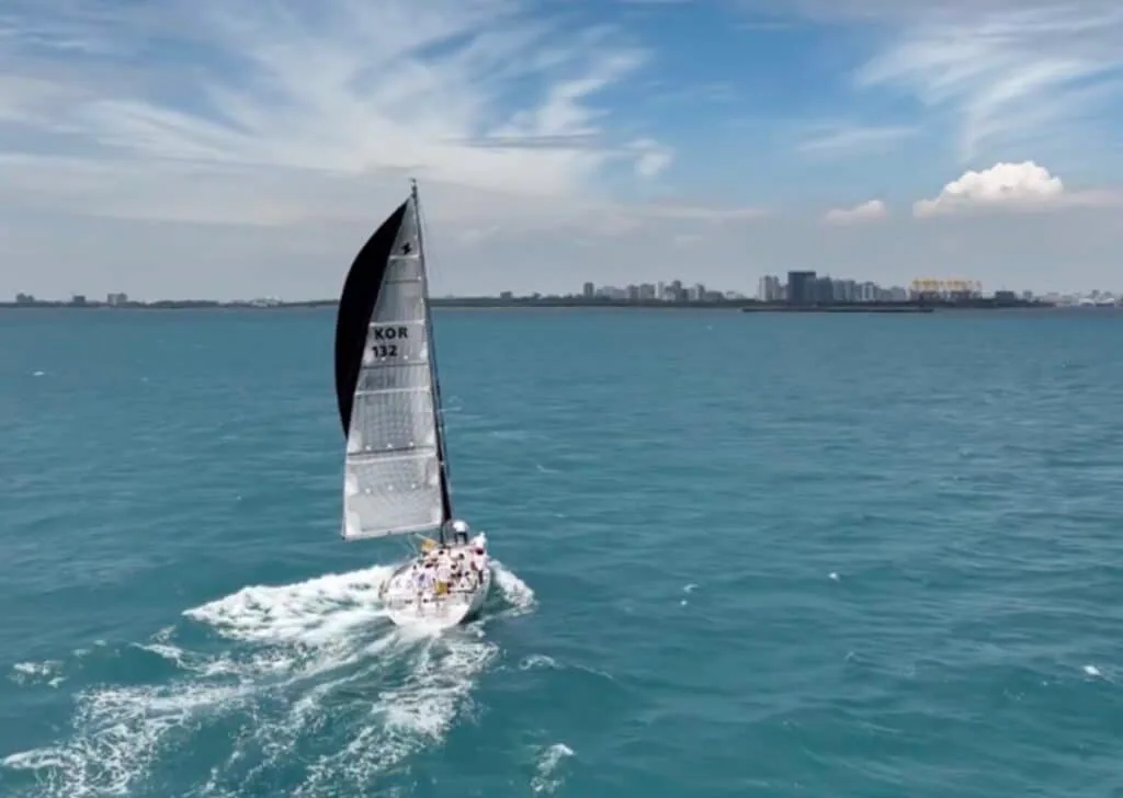 Yacht with a black-topped mainsail reaches past a coastal city skyline