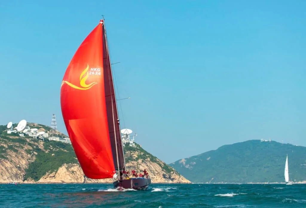 Yacht under a red spinnaker passes a hillside dotted with radar domes