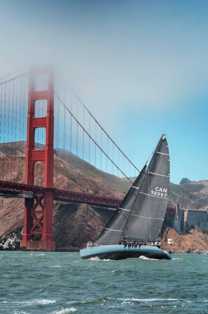Racing yacht CAN 18997 heeled under dark sails passing the fog-wrapped Golden Gate Bridge