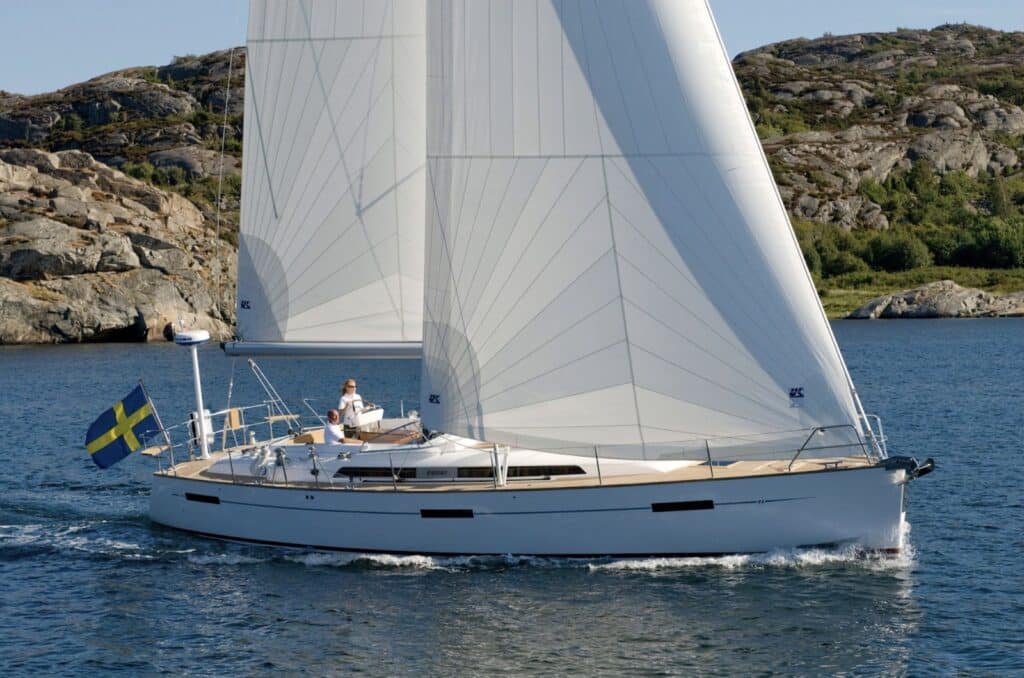 Cruising sloop flying a Swedish flag, sailing past a rocky coastline under white sails