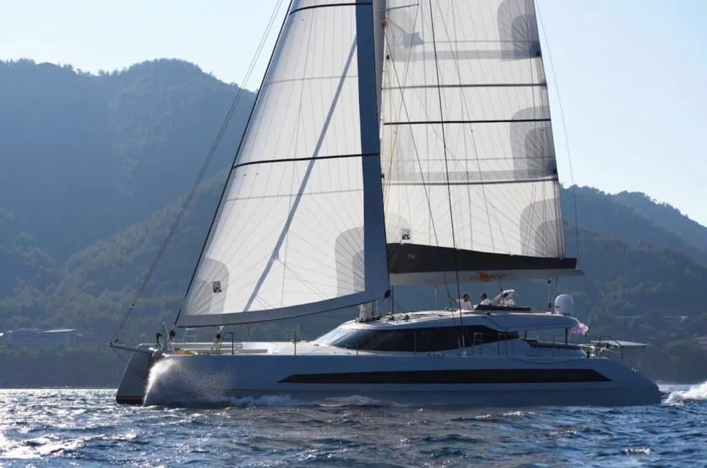 Cruising catamaran sailing past a mountainous coastline under white sails