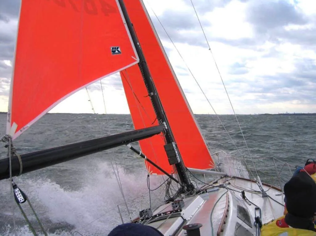 Yacht racing under bright orange-red sails in choppy water, crew braced in yellow gear