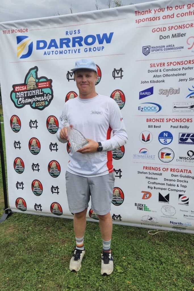 Sailor in a blue cap holding a glass trophy at the MCSA National Championship banner