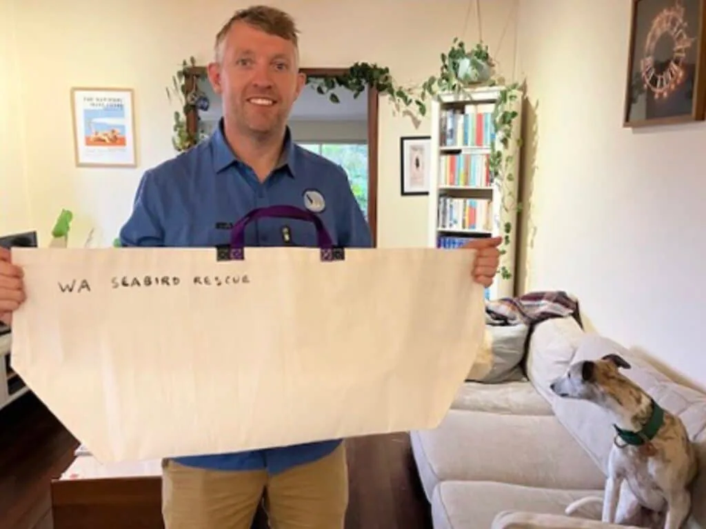 Man holding a recycled-sailcloth tote printed WA Seabird Rescue, with a dog looking on
