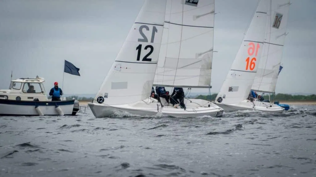 Team-racing keelboats numbered 12 and 16 rounding a mark past the committee boat