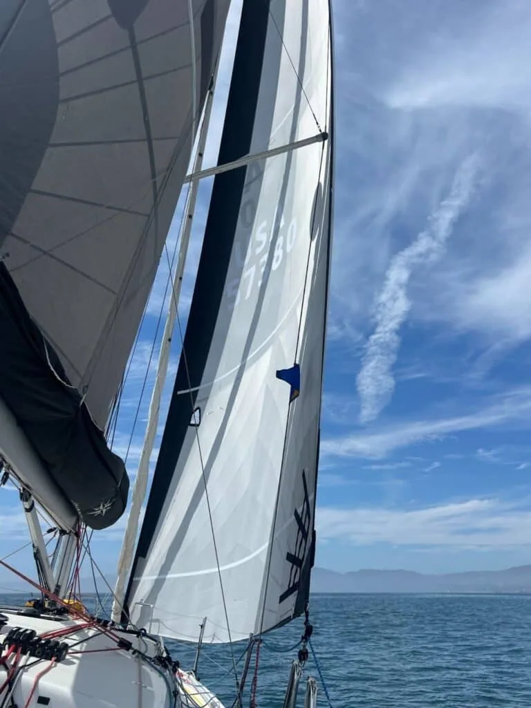 Deck-level view looking up a mainsail and genoa with hazy coastal hills beyond