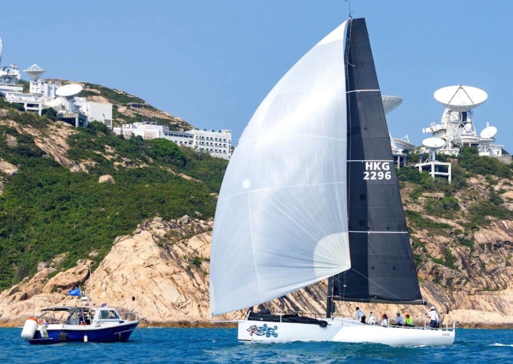 Yacht HKG 2296 reaching under spinnaker past a rocky headland with satellite dishes