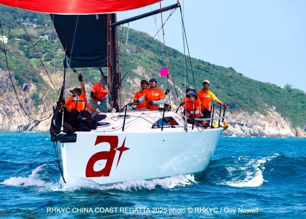 Crew in orange gear racing a boat marked a+ at the China Coast Regatta, hills behind