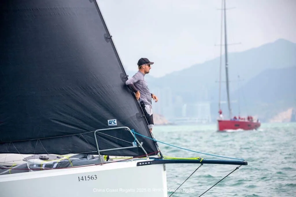 Sailor balanced on the hiking rack of a multihull, dark mainsail catching the wind, hills behind