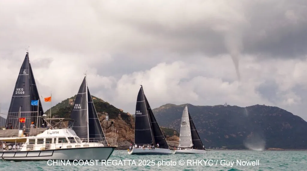 Dark-sailed fleet racing past a headland with a waterspout forming offshore