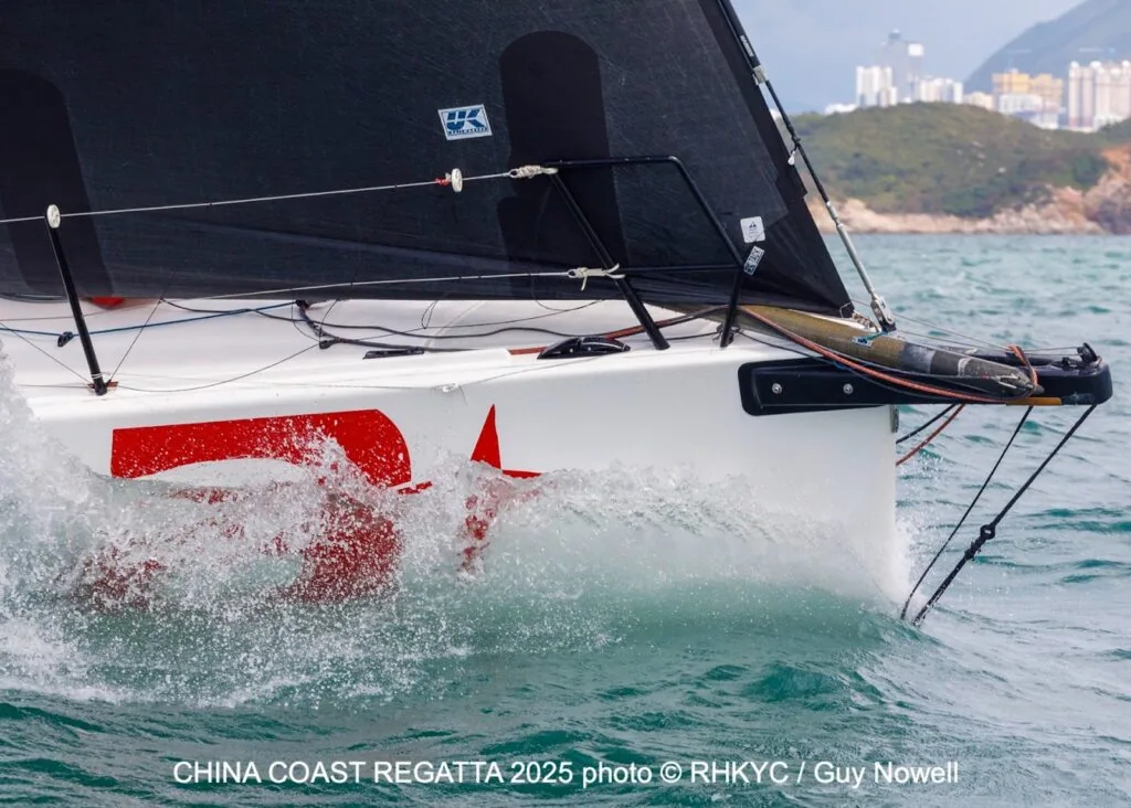 Bow-on view of a red-hulled racer crashing through waves, Hong Kong skyline behind