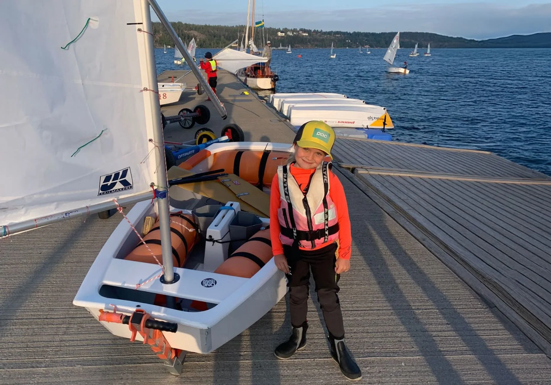 Young sailor in a life jacket beside an Optimist dinghy on the dock at sunset