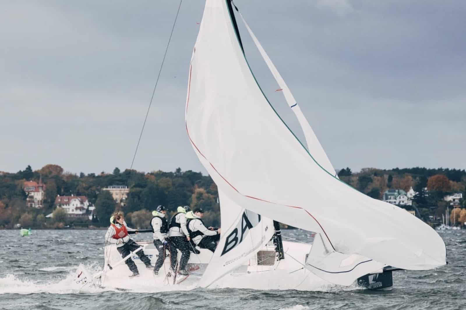 Crew hiked out hard on a heeling keelboat during a close race, houses lining the shore behind