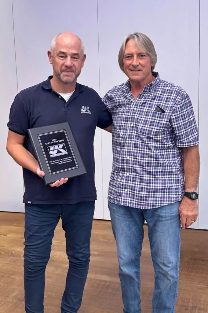 Two men posing indoors with an engraved UK Sailmakers award plaque