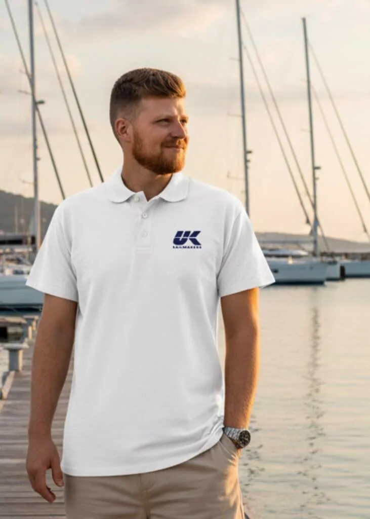 Man wearing a white UK Sailmakers logo polo shirt on a marina dock at sunset