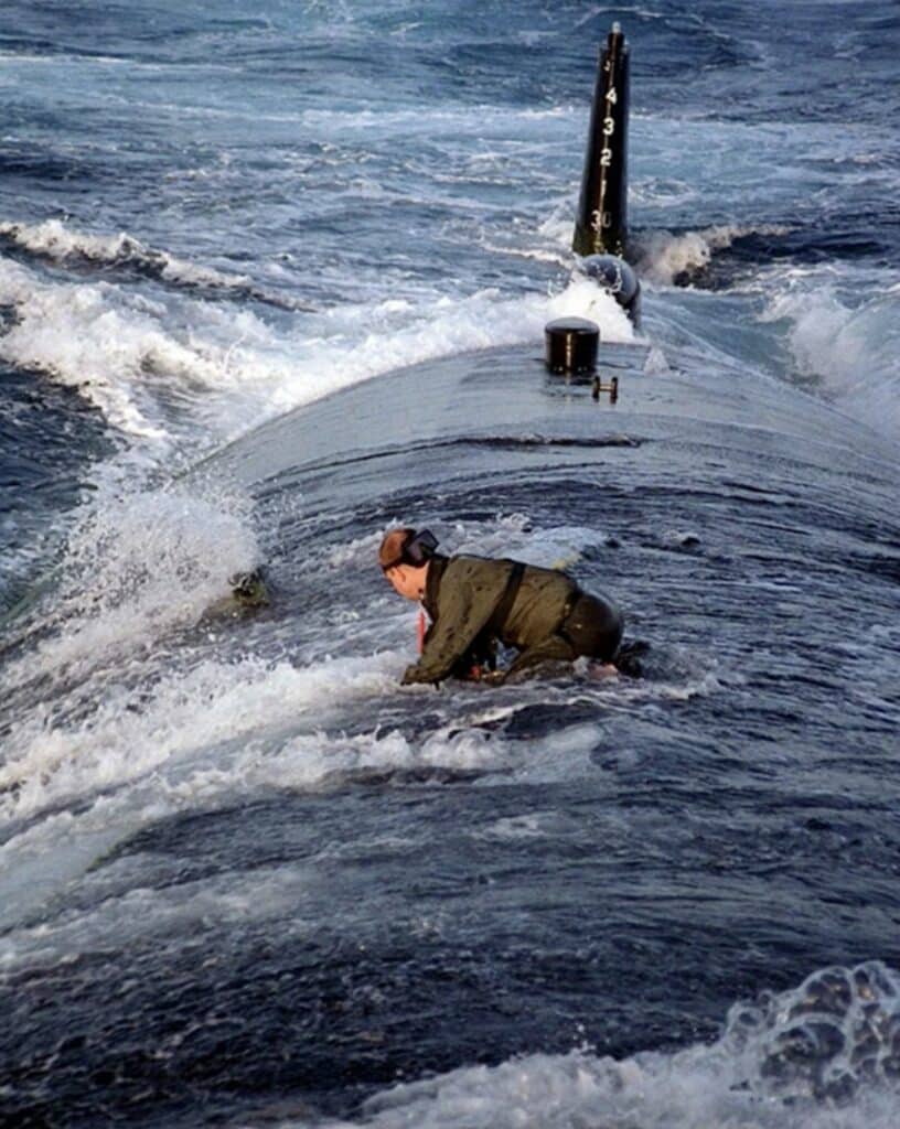 Navy sailor braced on a submarine's deck as waves wash over the hull