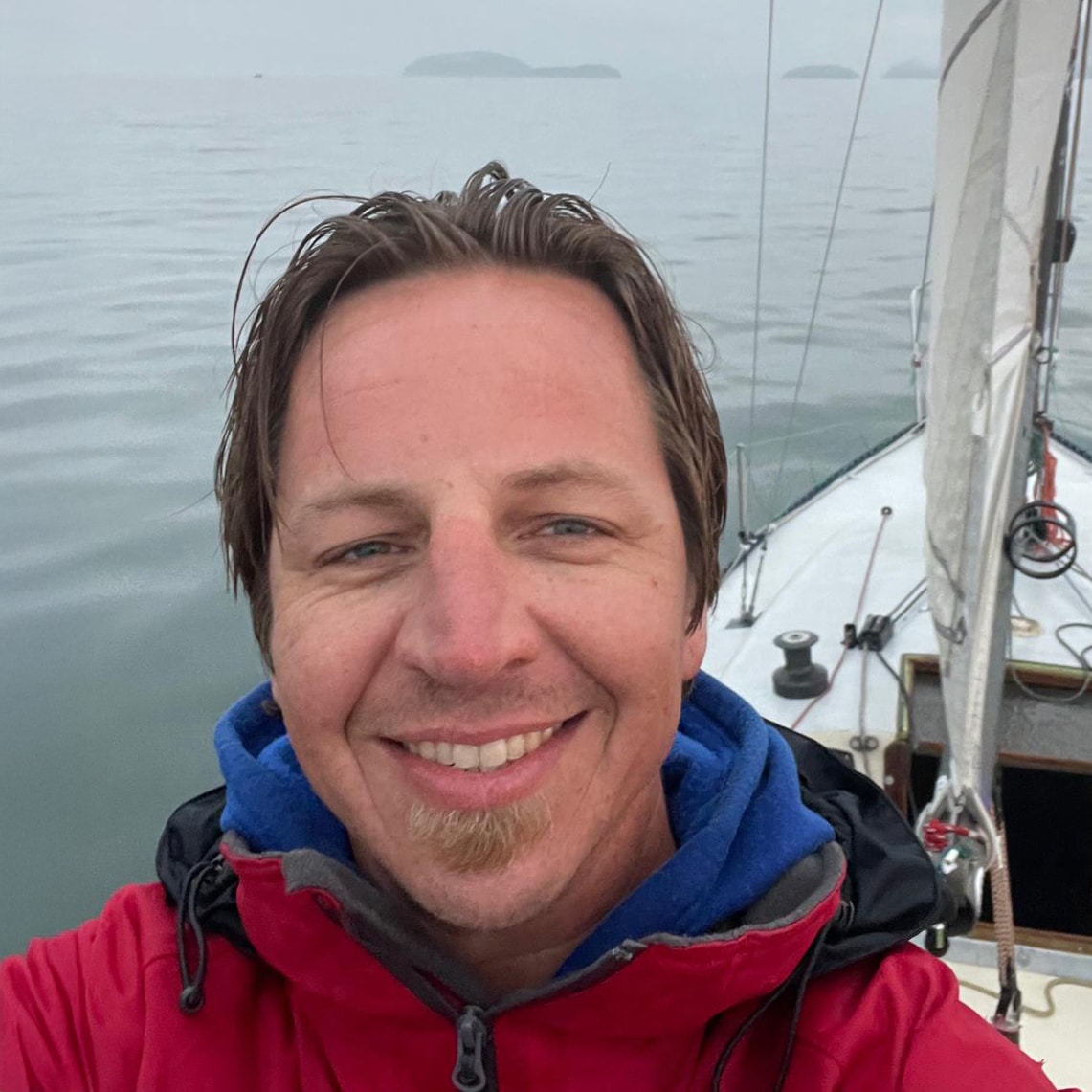 Selfie portrait of a man in a red jacket on a sailboat under overcast sky