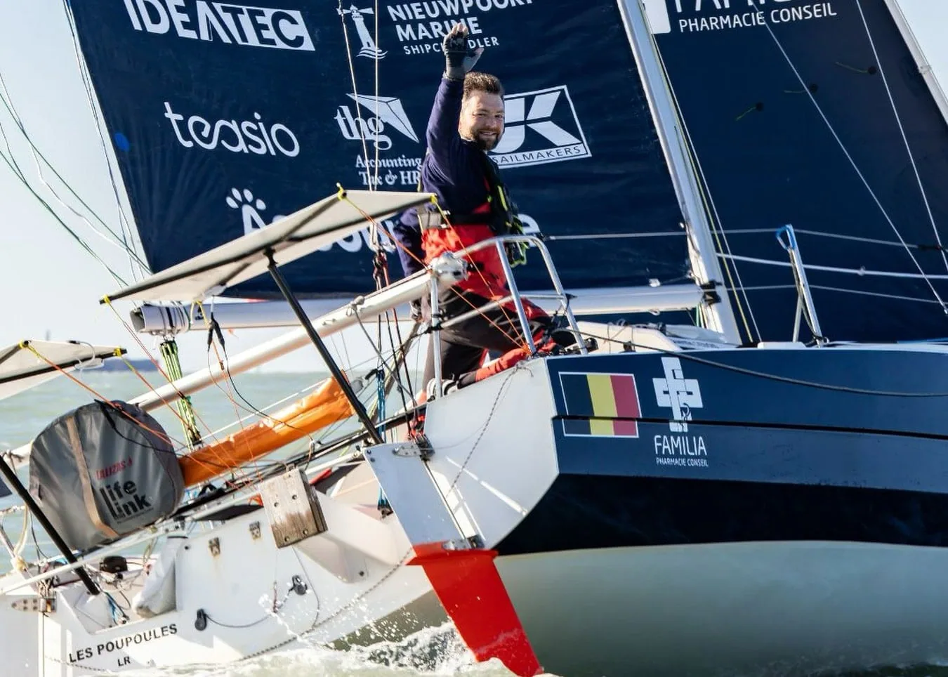 Skipper waving from ODIGO's deck under her sponsor mainsail, Belgian flag on the hull