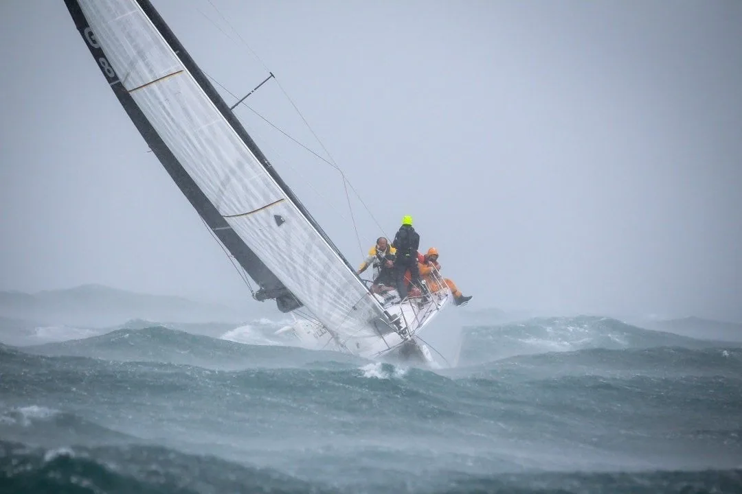 Sailboat heeled hard in grey rough seas, crew hiked out in foul gear