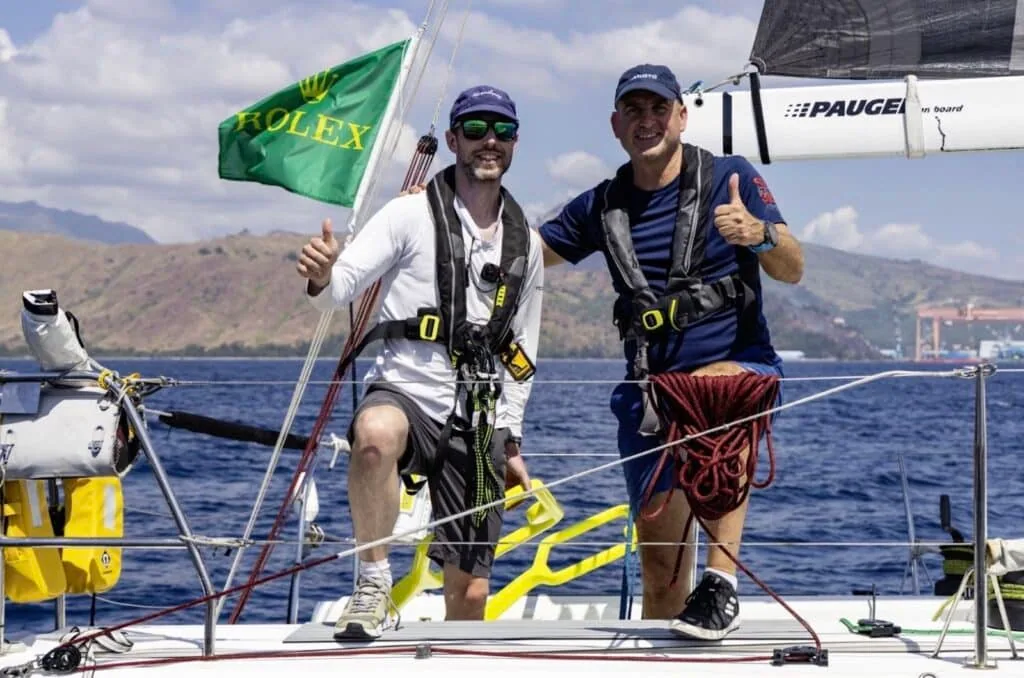 Two crew giving thumbs up on deck with a green Rolex flag flying