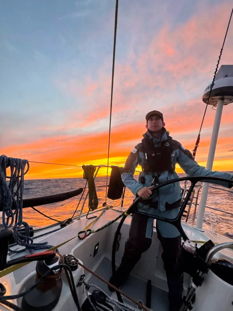 Sailor at the wheel under a dramatic orange sunset offshore