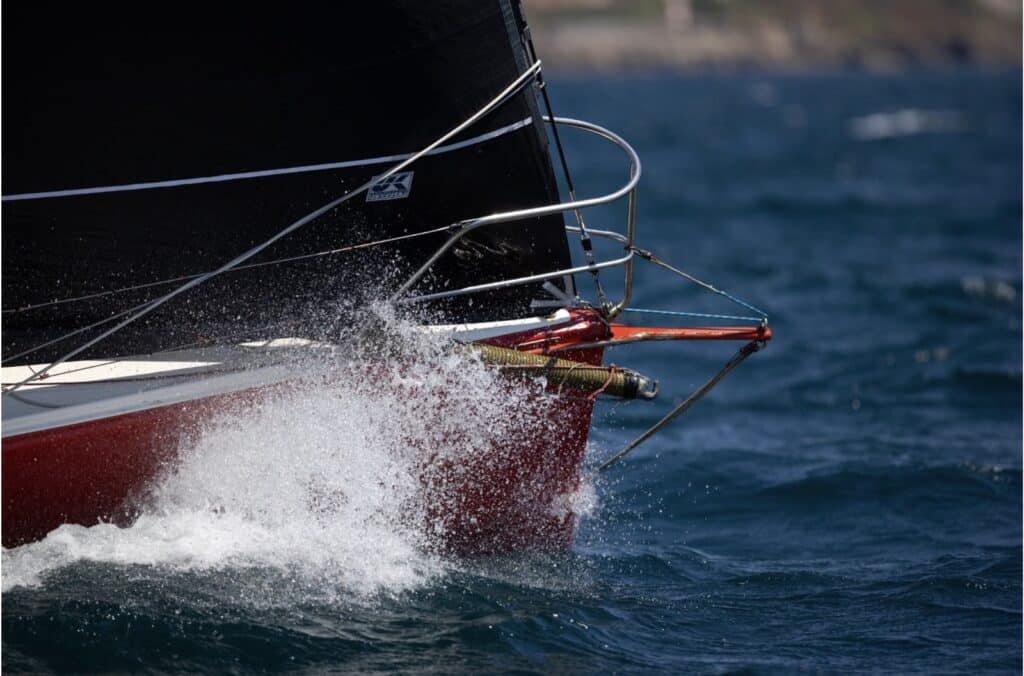Close-up of a red hull's bow throwing spray under a black jib