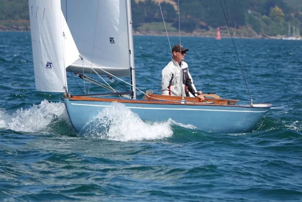 Helmsman in a blue-hulled classic day boat under white UK Sailmakers sails, spray flying