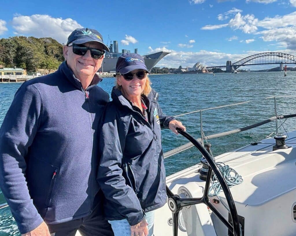 Couple at a sailboat's helm in Sydney Harbour, with the Harbour Bridge and Opera House behind