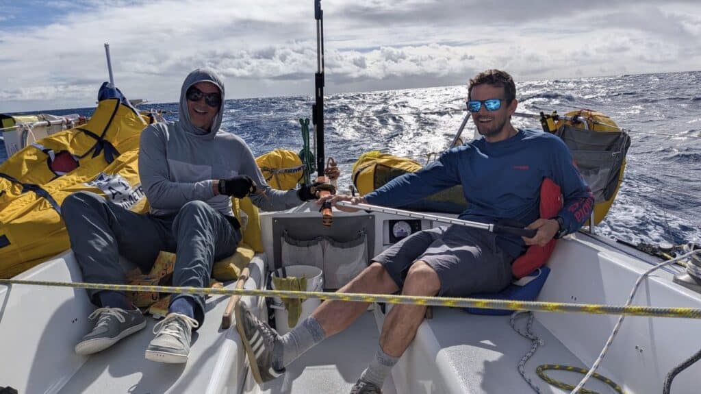 Two Dorado crew relaxing in the cockpit mid-Pacific, yellow sail bags stacked on deck