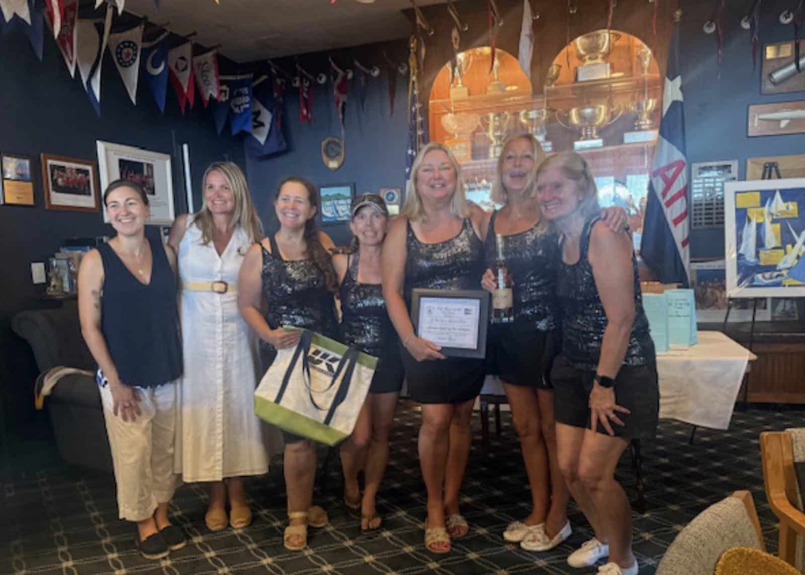 Seven women of a winning crew pose with their trophy and a UK Sailmakers bag in a yacht club trophy room