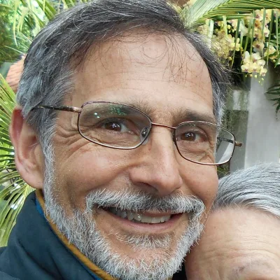 Adam Loory, smiling, with a grey beard and wire-rimmed glasses, photographed outdoors among greenery