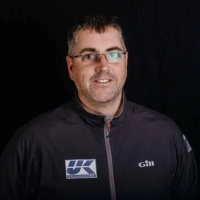Barry Hayes in glasses and a dark UK Sailmakers jacket, smiling against a black studio background