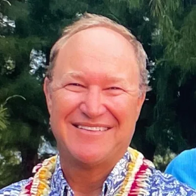 Brendan Huffman smiling in a blue floral shirt and flower lei, with trees in the background