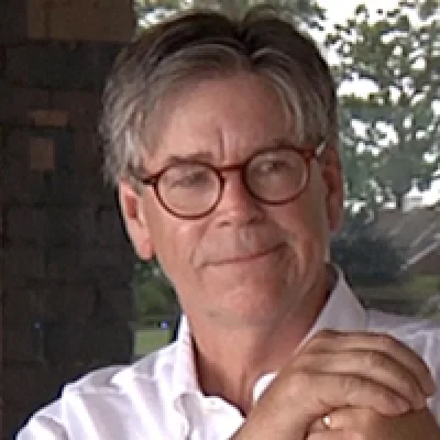 Buttons Padin in round tortoiseshell glasses and a white shirt, seated on a porch with trees behind