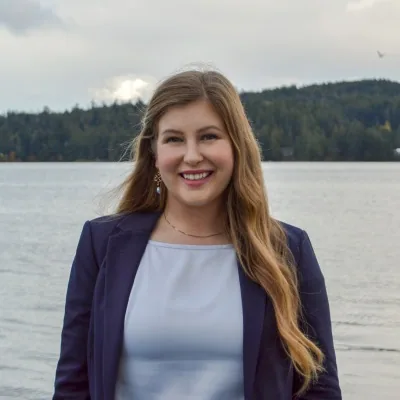 Heather Mahady in a navy blazer, smiling at a waterfront with a forested shoreline behind her