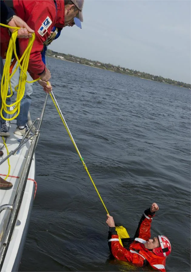Crew in a red jacket reaches a pole and yellow line down to a swimmer in an orange survival suit alongside