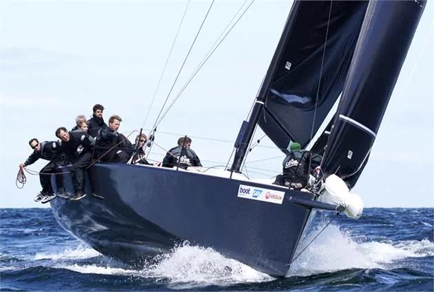 Dark-hulled race yacht hard on the wind under black Uni-Titanium sails, crew hiking on the rail through a bow wave