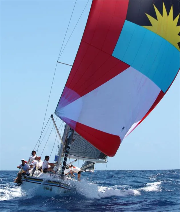 Yacht running under a red, white and blue spinnaker printed with the Antigua flag, crew on the foredeck