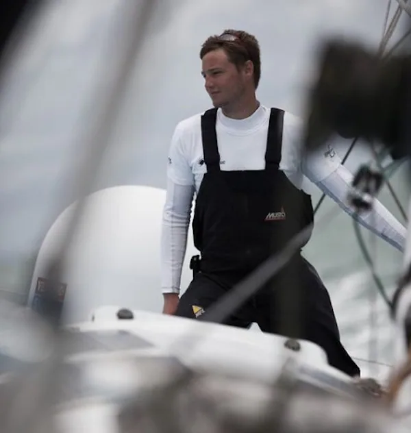 Oscar Mead on deck in a white top and black sailing salopettes, framed by rigging