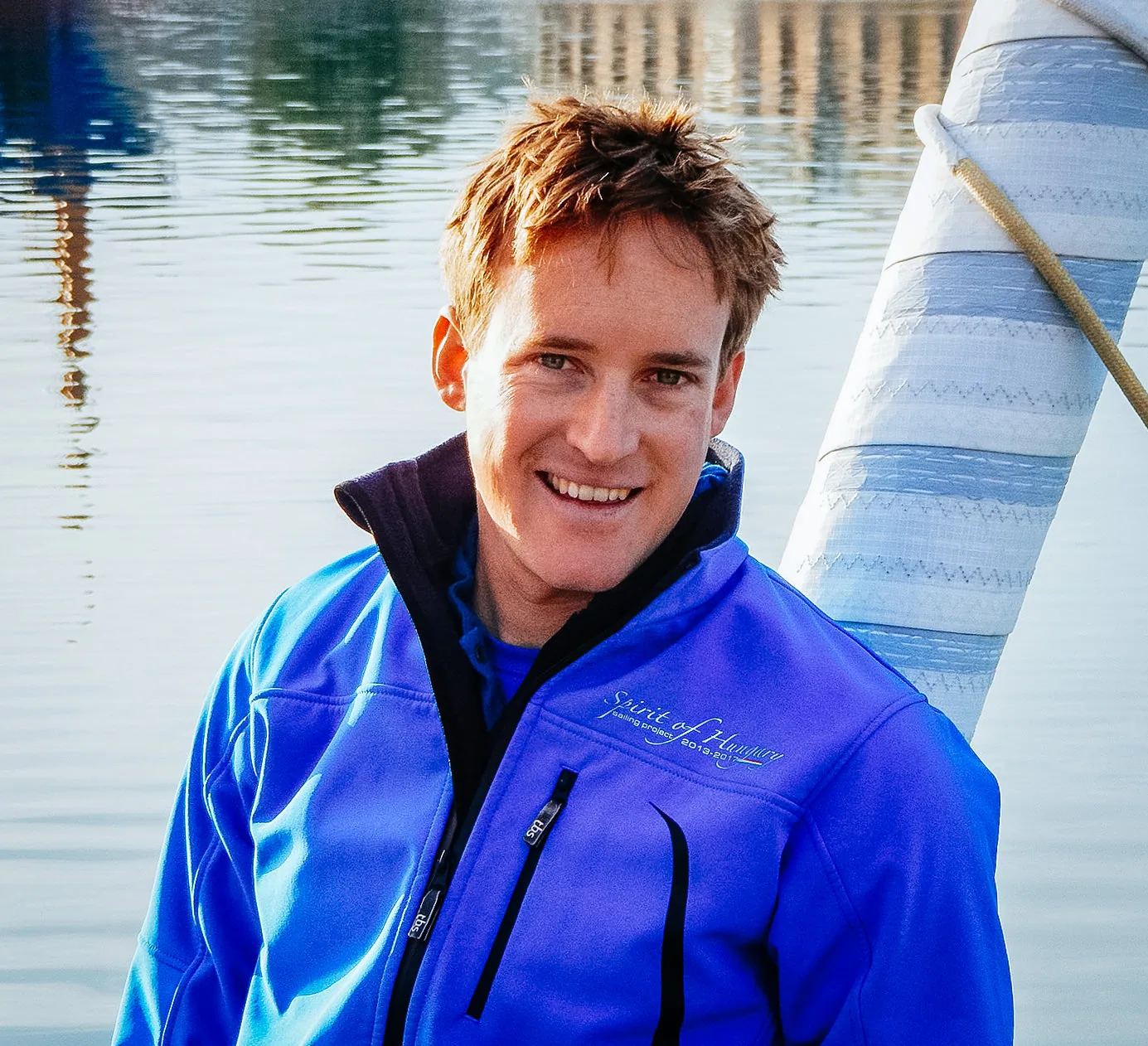Smiling young sailor in a blue Spirit of Hungary jacket beside a furled sail at the dockside