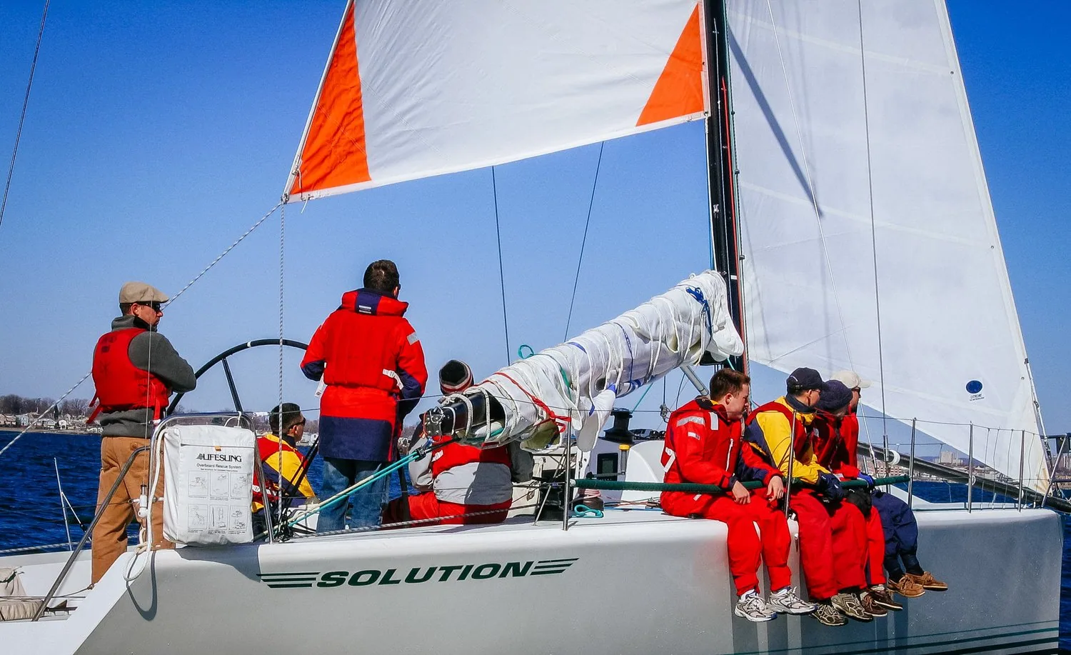 Orange-cornered storm trysail set on SOLUTION with the main furled on the boom, crew in red jackets on the rail