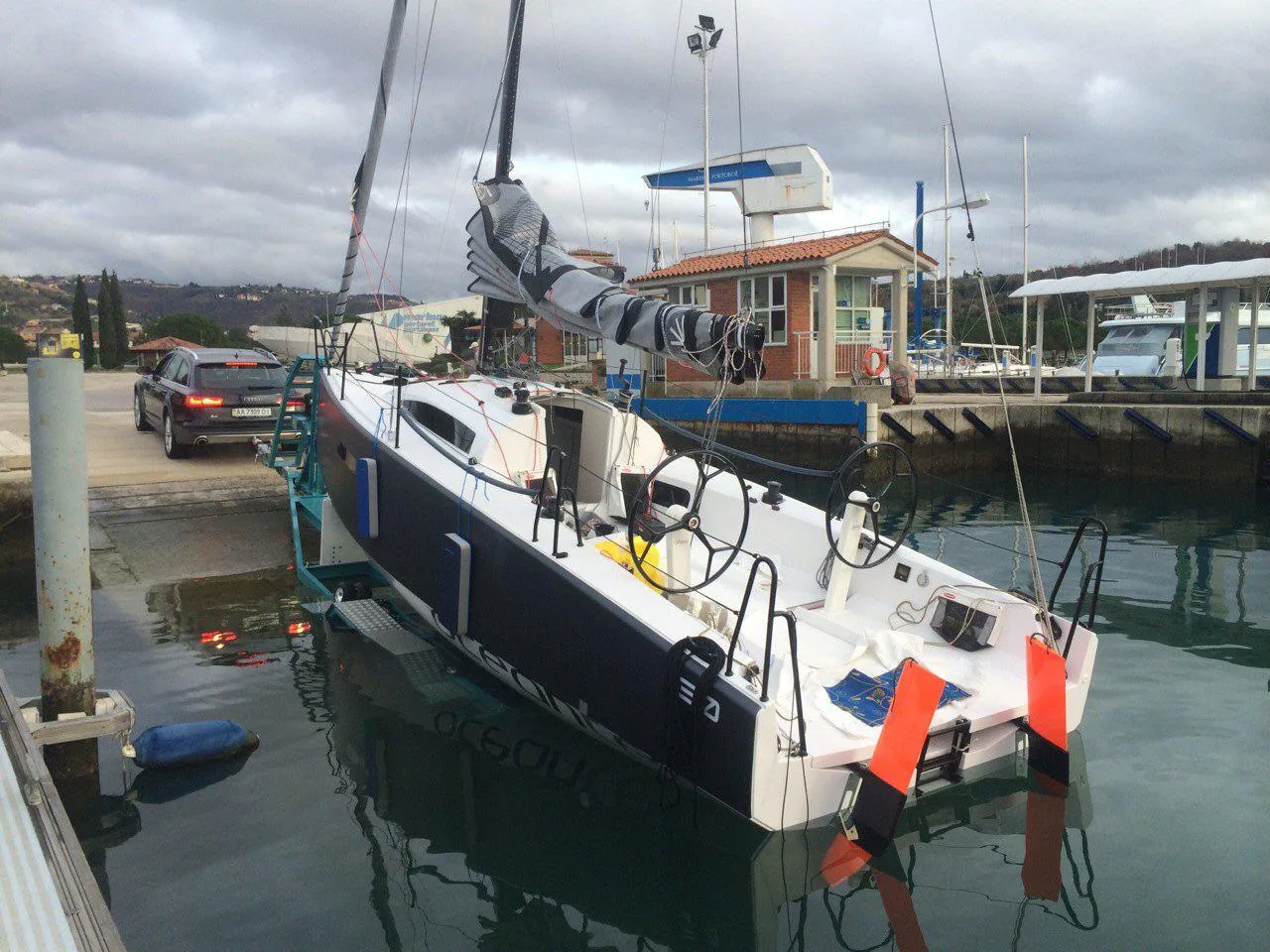 Luka 30 with a dark blue bow afloat at a marina slipway, mainsail flaked on the boom and fenders out