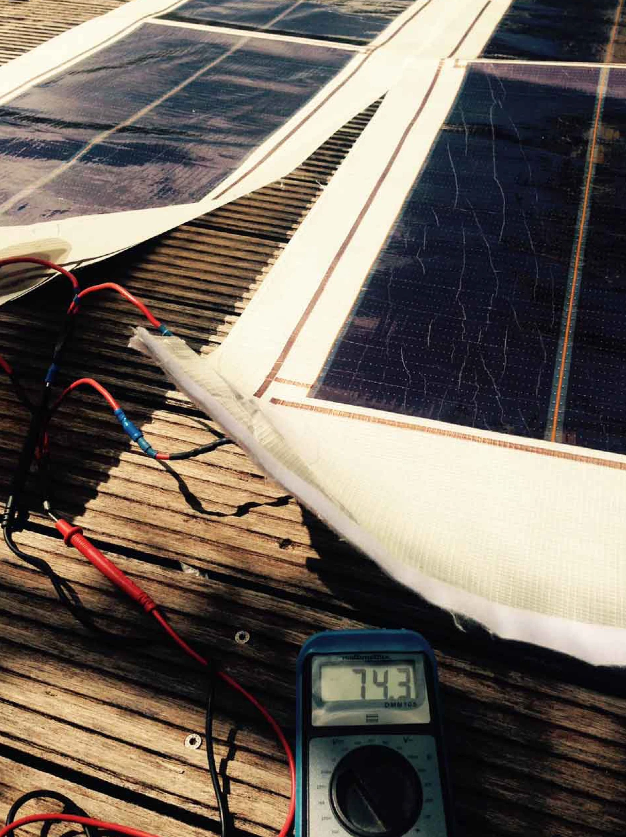 Thin-film solar strips laminated into sail panels on a wooden deck, wired to a multimeter reading 74.3
