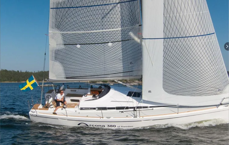 Arcona 380 sailing under grey laminate mainsail and genoa, Swedish flag at the stern, wooded shore behind