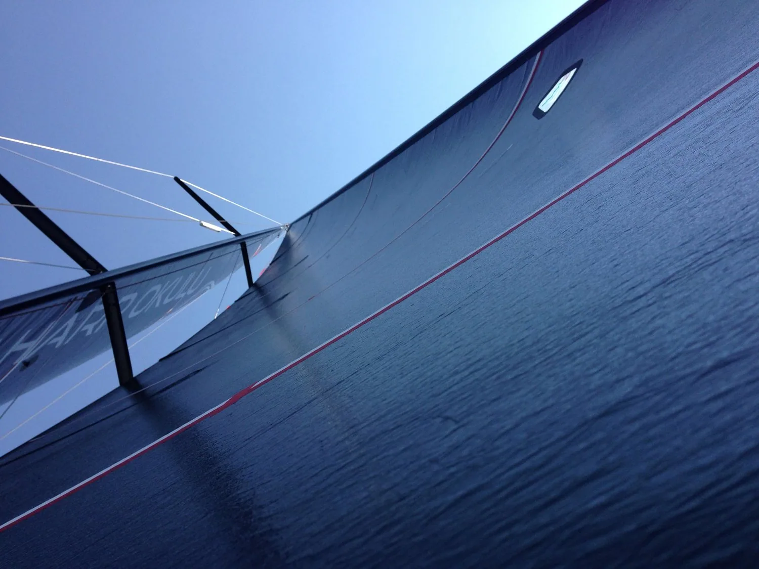 Looking up a black laminate mainsail from the deck, red draft stripe running across it under a clear blue sky