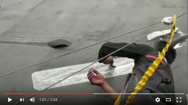 Video still: crew member pressing a clear patch onto a grey Volvo 70 mainsail on deck while underway