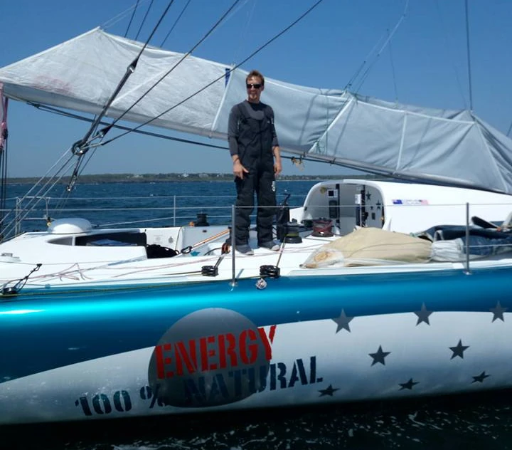 Conrad Colman standing on deck, mainsail flaked on the boom, 'Energy 100% Natural' and stars on the blue hull